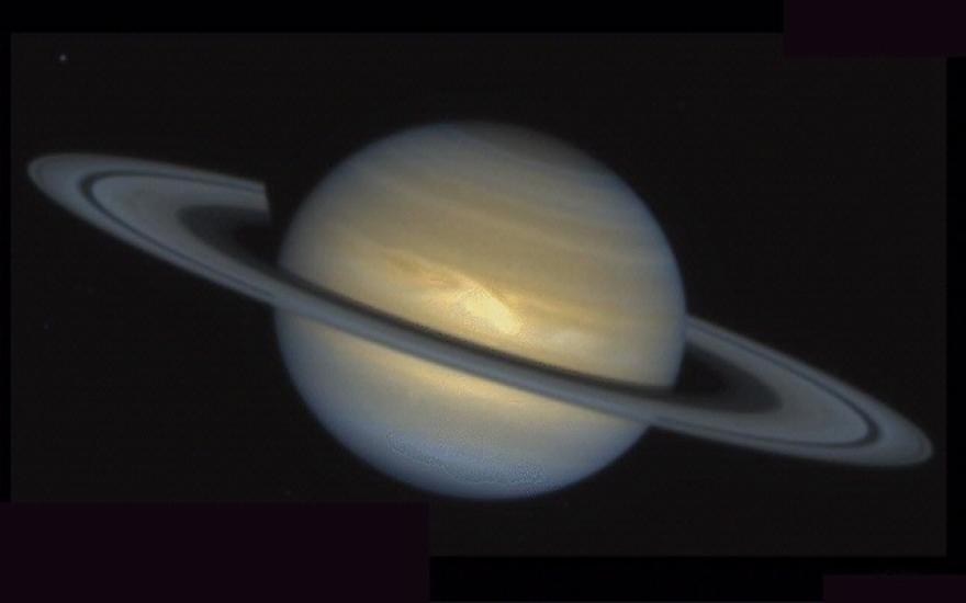 Disk view of Saturn with a large storm appearing as lighter colors near the center of the planet above the rings.