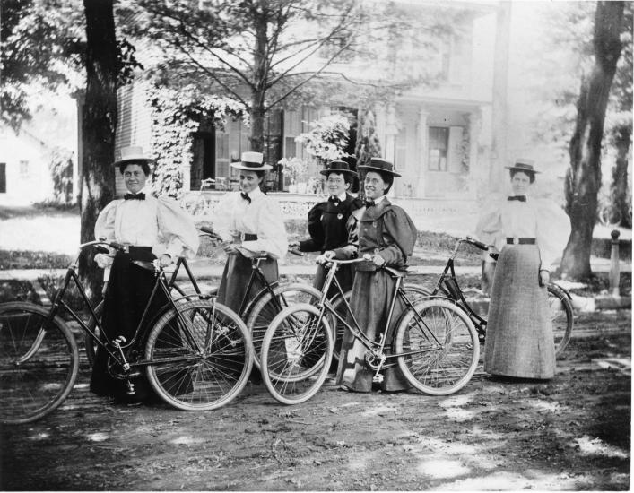 A group of five women in 1900s style dress with bicycles. 