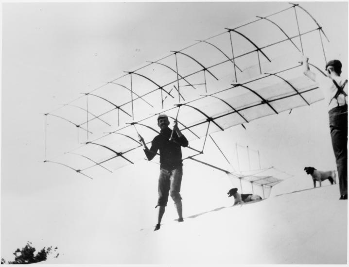 A man attached to a simple glider with two dogs behind him and a man holding up the glider on the right side.