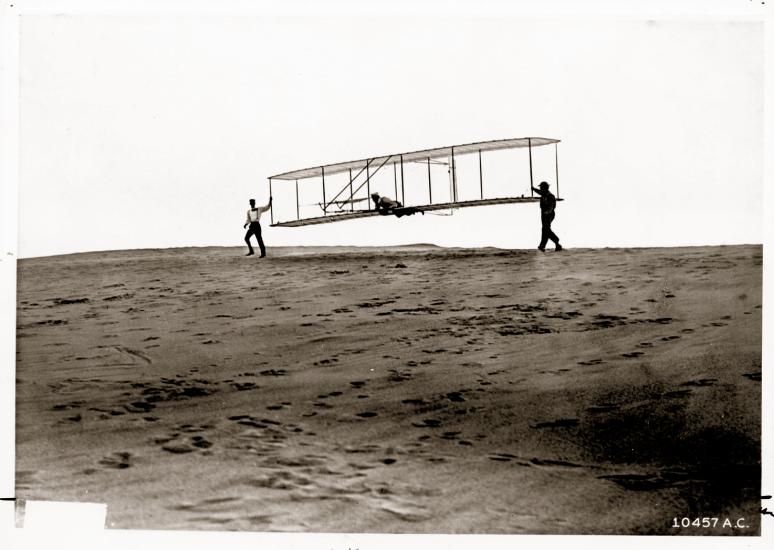 Orville Wright is prepared for takeoff in a test using the 1902 Wright Glider. The Wright Glider is being carried by Wilbur Wright and Dan Tate.