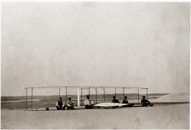 Six men sit on the ground in front of a glider