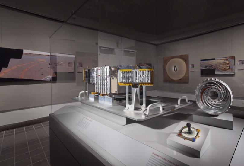 Multiple parts of the Rover robotic unit inside a display celebrating the Rovers. An electronic module made of multiple metal pieces can be seen on the left. On the right side, a cylindrical prism with one open side represents another part of the Rover.