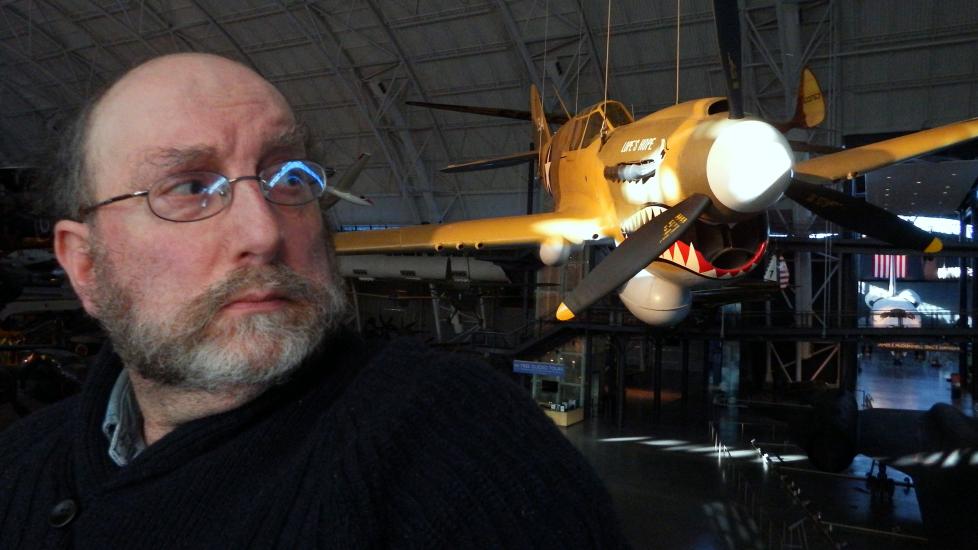 A white male specialist at the museum poses in front of a yellow monoplane with a shark mouth painted on the lower nose.