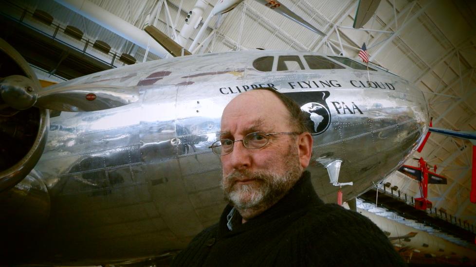 A white male archives specialist at the museum poses below a silver commercial airplane.