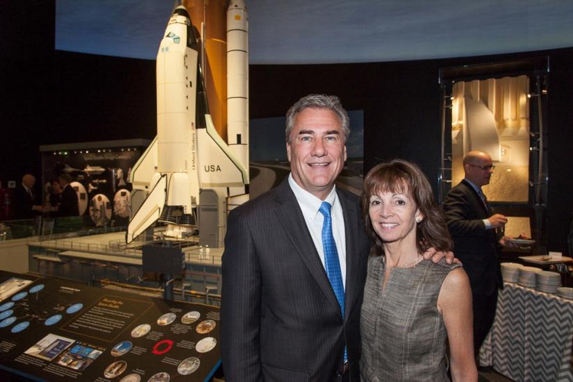 Two people, a white male and a white woman, pose together with a backdrop of a space shuttle model behind them.