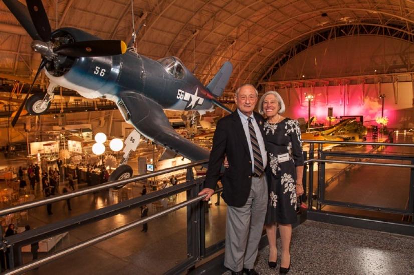 Robert and Lenore Briskman, a couple consisting of a white man and woman, pose together with the backdrop of a gray and white monoplane behind them.