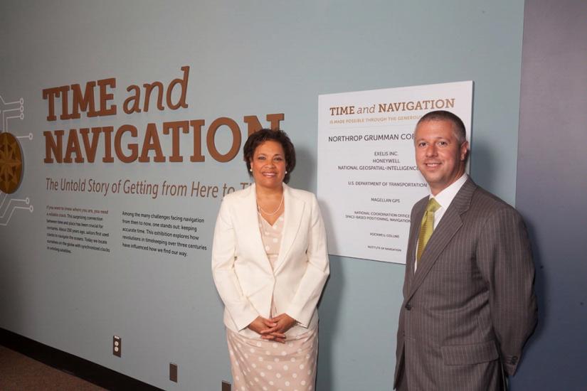 Two people, a woman and a male, stand next to the sponsorship plague for an exhibit that their corporation sponsored.