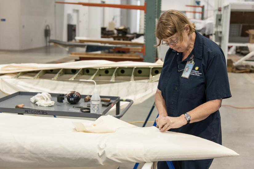 A white female museum specialist performs restoration work involving fabric on the Curtiss Helldiver, an aircraft.