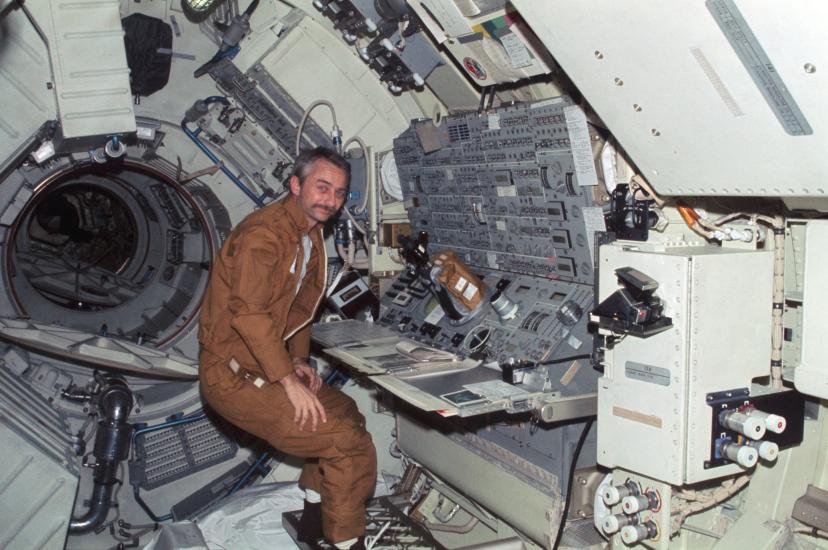 Owen Garriott, a male astronaut, stands near a console inside a space station.