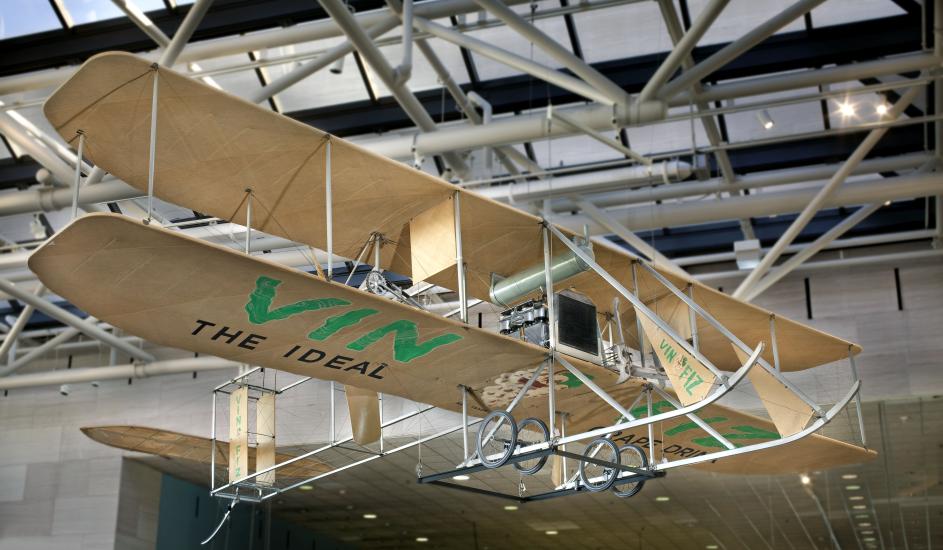 Biplane made with tan fabric and white metal hanging in the museum. "Vin Fiz" written on the bottom part of the lower wing in green paint.