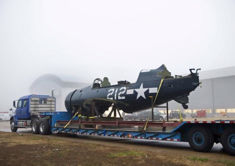 The fuselage of a blue military monoplane is transported on a blue truck between Museum facilities.