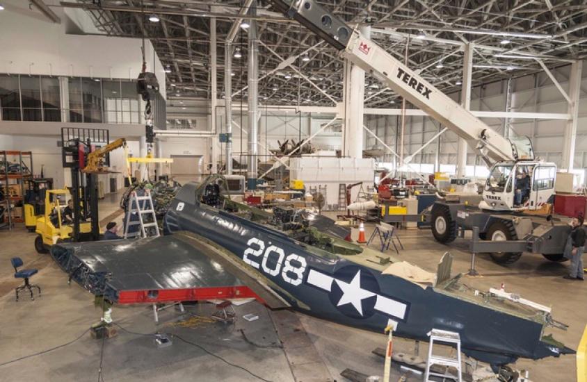 The engine of a blue military monoplane is placed by crane into the aircraft.