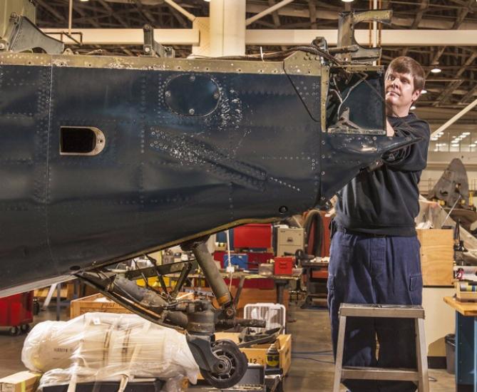 A museum specialist examines the tail and rudder of a blue military monoplane.