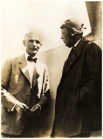 Charles Lindbergh and Henry Ford, both of whom are white men, stand together. Lindbergh is wearing aviator gear.