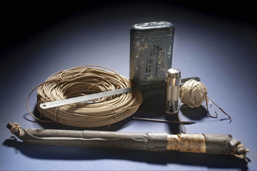 A set of emergency survival suplies including a fishing line and hook, a silver-colored blade, an emergency flare, a tan-colored roll of cord, an a metal container of emergency rations.