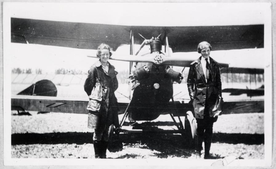 Neta Snook, a female flight instructor, stands with her at-the-time female pilot student, Amelia Earhart, in front of a biplane with one engine.