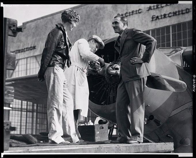 Paul Mantz, a man, stands to the right of Amelia Earhart, a white woman, as they speak to each other.