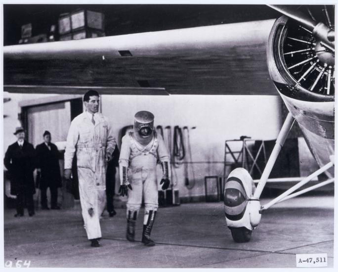 Wiley Post walks under his monoplane, wearing a pressure suit he planned to test while flying his aircraft. The pressure suit features a cylinder-shaped helmet and dark-colored boots.