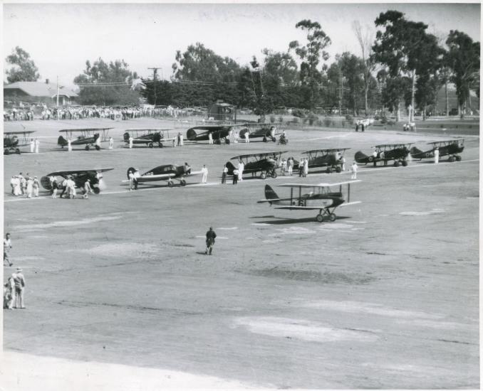 Two lines of early aircraft, mostly biplanes, are seen as part of an air race featuring only women pilots.