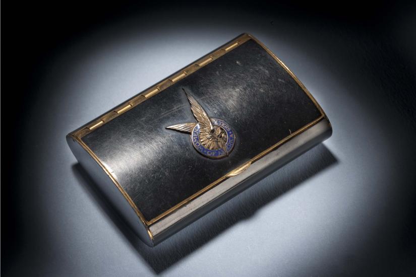 Black makeup case with closed lid. On the center of the lid, there is a silver insignia of a pair of two wings inside a blue seal.