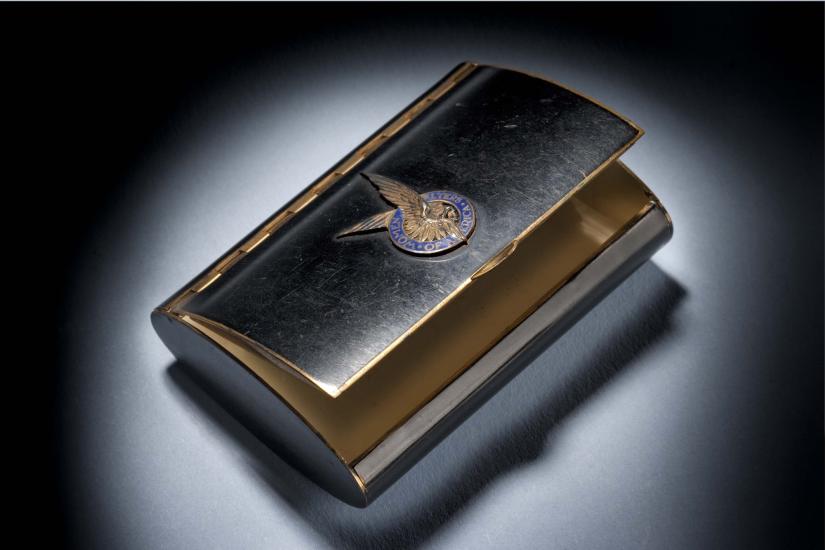 Black makeup case with open lid. On the center of the lid, there is a silver insignia of a pair of two wings inside a blue seal.