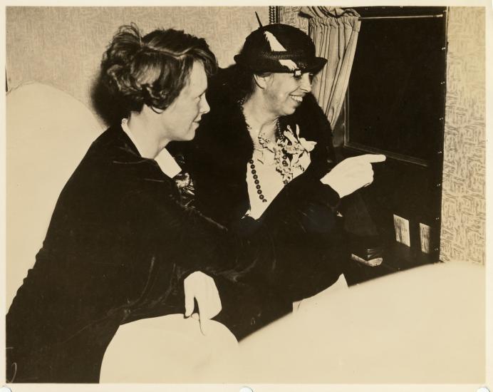 Amelia Earhart and Eleanor Roosevelt, both of whom are white women, sit and look at something not visible.