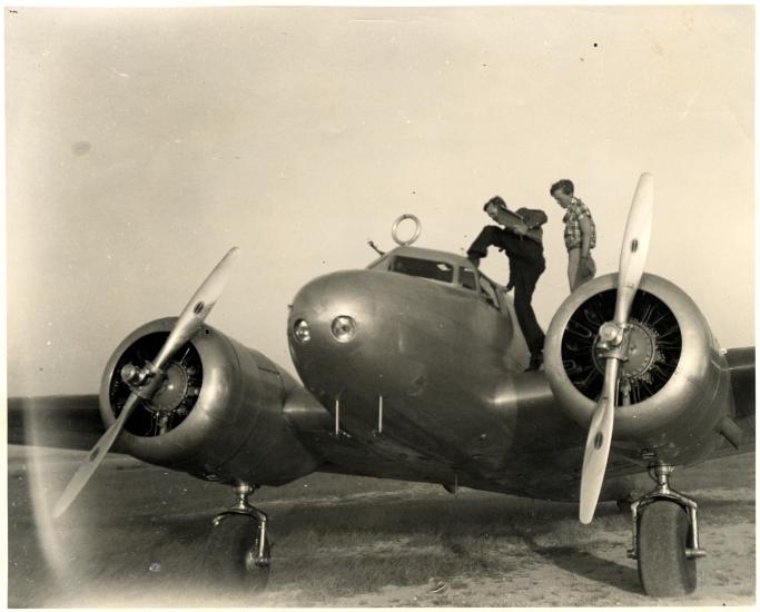 Amelia Earhart and Fred Noonan, a white woman and white male respectively, stand on top of a monoplane with twin engines. Noonan is climbing into the monoplane.