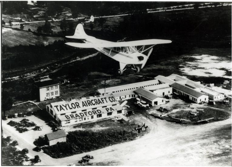 A monoplane flies over a set of buildings which are labeled as belonging to the "Taylor Aircraft Co." on the roof of a building on the left of the other buildings. Below the roof stating the company's name is the location of "Bradford, PA" on a lower roof.