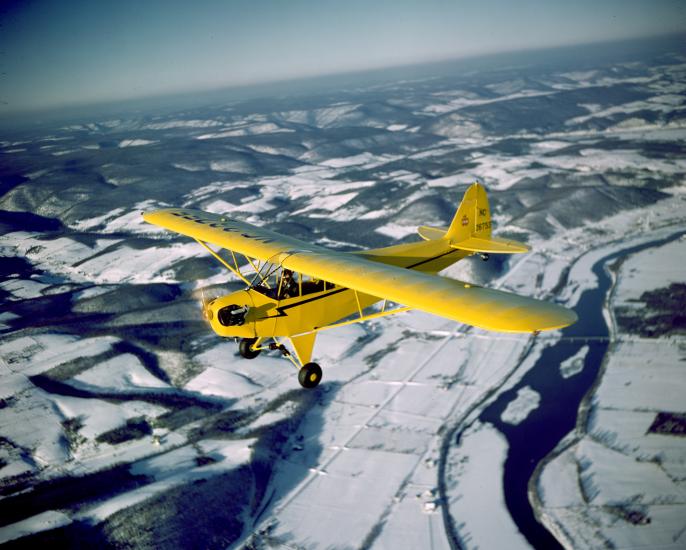 A yellow monoplane with one engine flies above land.