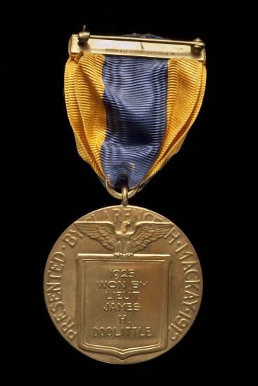 Back of aviation medal. Medallion features an etching of an eagle holding a plaque stating the winner of the medal and the rounded edges state the presenter of the medal. Suspended using yellow and black fabric attached to metal pin.