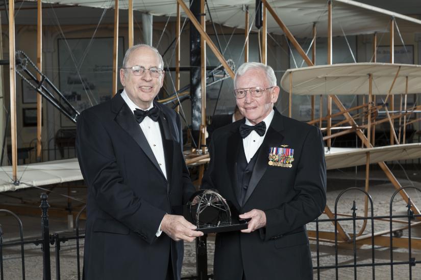 Two white men stand together, holding a trophy award at a Museum awards event.