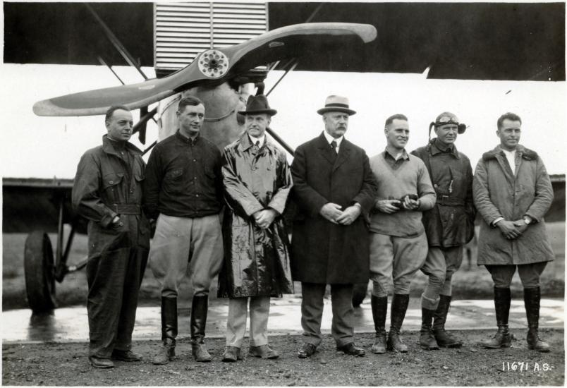 U.S. President Calvin Coolidge (a white man wearing a raincoat) stands with other white people at a meeting with pilots.