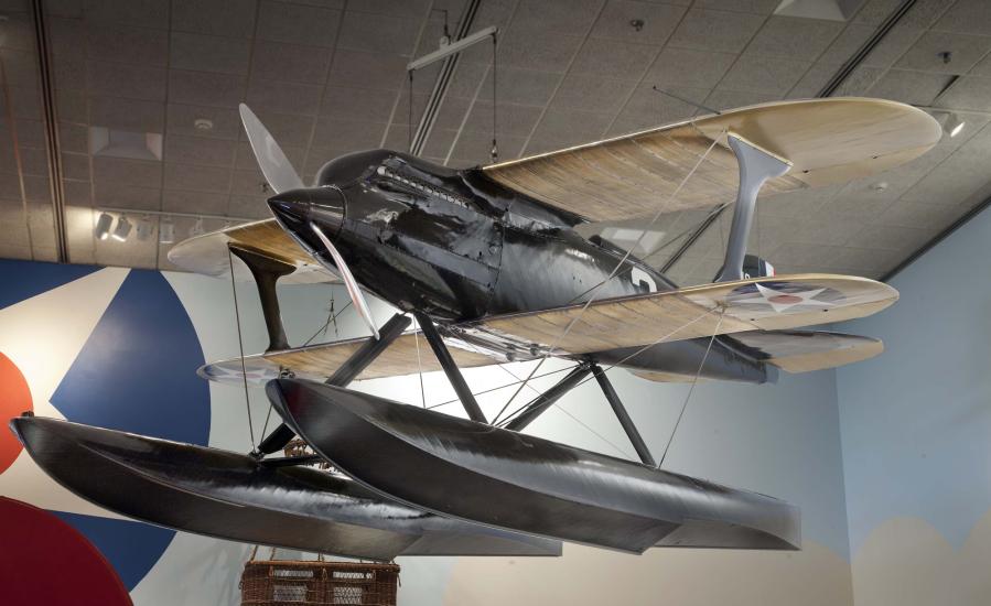 Diagonal and frontal view of black biplane with single engine and pontoon floats hanging in the Museum.