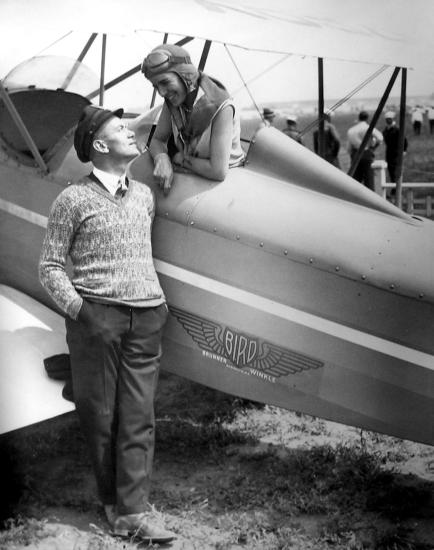 A woman in a pilot's cap leans out of a plane. A man standing on the ground stares up at her. The photo is in black and white.