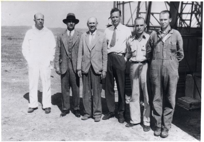 Six white men, including Harry Guggenheim, Robert Goddard, and Charles Lindbergh, stand in front of a tower in a desert.