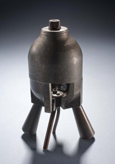 Metal rocket motor with four leg-like tubes below a cylinder-shaped combustion chamber. Small cutaway revealing the interior of the chamber.