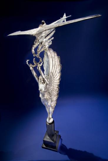 Silver-colored trophy with topper of a winged angel holding olive wreaths which are picked up by a plane.