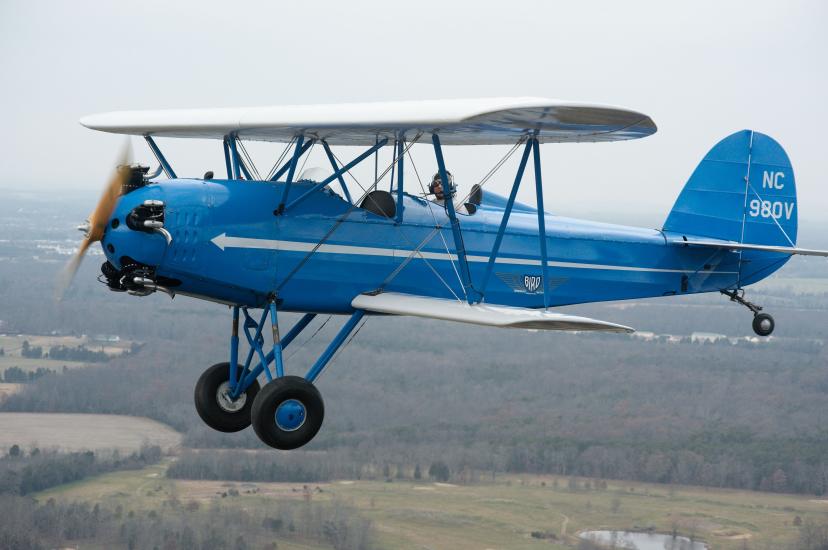 Side view of blue biplane with white wings, one engine, and fixed landing gear. Biplane is in flight. Registration number "N980V" painted in white paint on rudder.