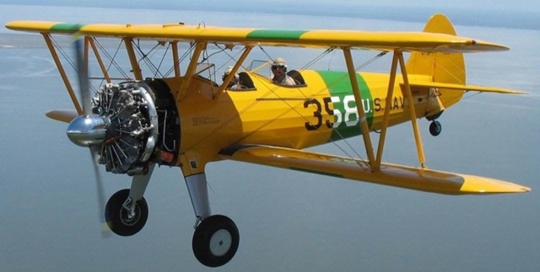 Side view of yellow and green biplane with single engine and fixed landing gear. Biplane is in flight.