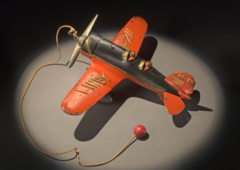 Top view of orange and black monoplane toy with a ball and string attached to the center of the propellors on the front.