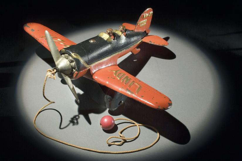 Top view of orange and black monoplane toy with a ball and string attached to the center of the propellors on the front.