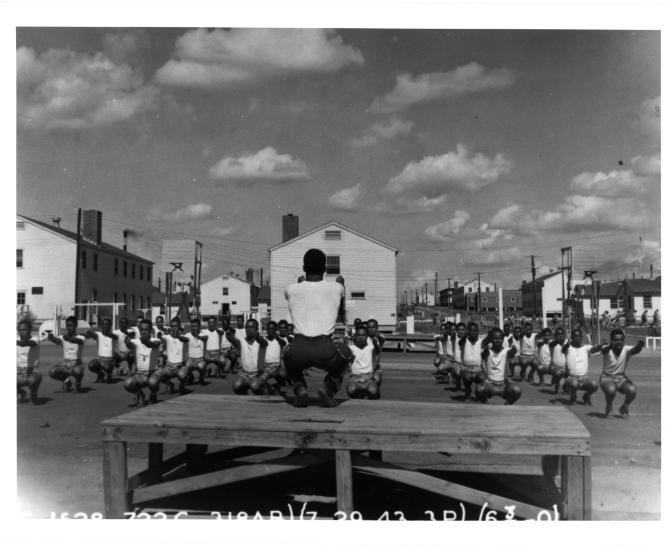 Partial view of some of the buildings and a clearing at the Tuskegee Army Air Field. A person runs physical training for cadets in the foreground.