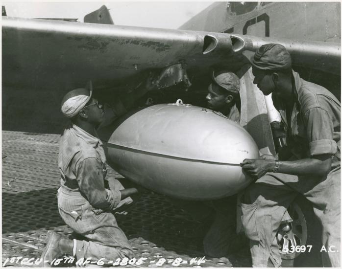 Members of a ground crew for a U.S. fighter group install a fuel tank onto the wing of a fighter plane.