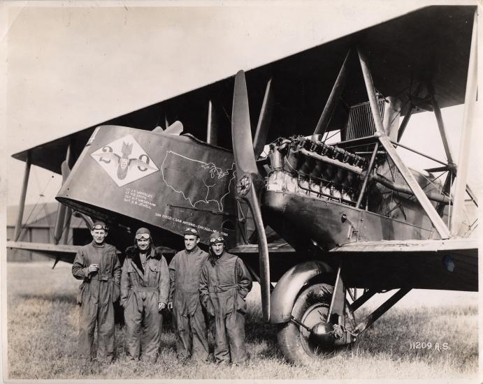 A group of white males representing the U.S. Army Air Service stand next to the nose of a biplane.