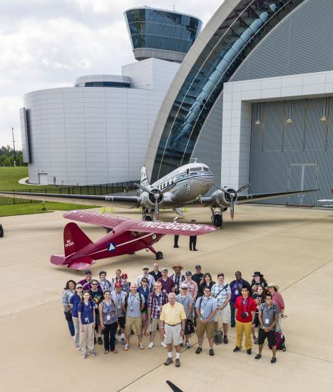 A group photo featuring several social media users who follow the Museum, the volunteers of an event, and the Museum's former director. They are standing on the runway in front of two aircraft.