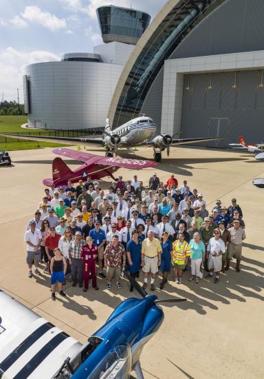 A group of pilots and crew members who participated in a pilot event at the Museum stand together with a former director of the Museum for a group photo.