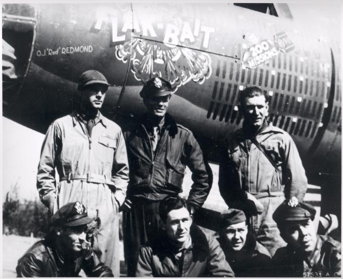 A group of seven members of a bomber combat plane used in World War II named "Flak-Bait" pose formally in front of the aircraft following a milestone mission.