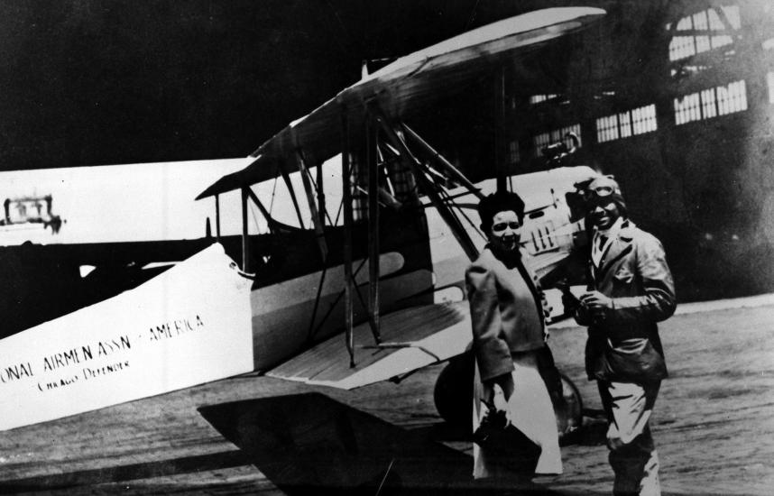 Chauncey Spencer, an African-American male pilot, stands next to a biplane with another person.