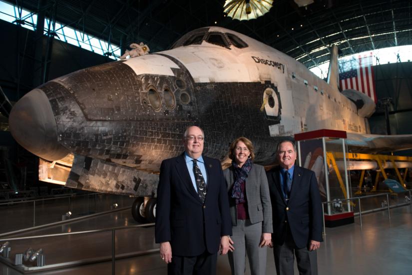 Three people, two white men and one white woman, stand formally in front of the Space Shuttle Discovery, a white spacecraft.