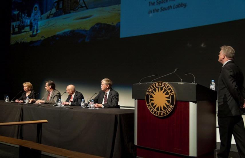 Five people present a lecture at the Museum. One is standing next to a podium whereas four other people are sitting at a table.